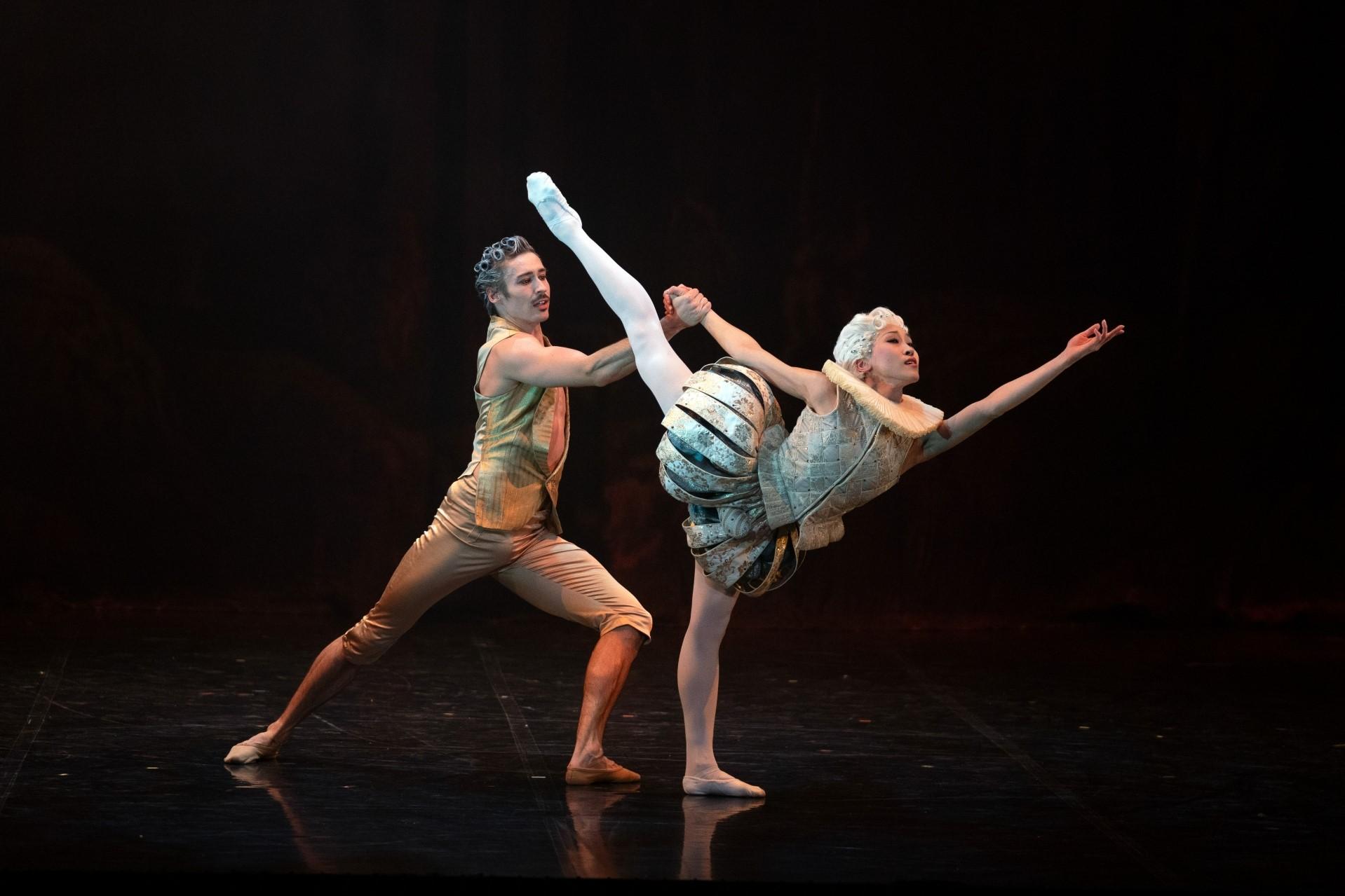 A female dancer in light, 18th century costume reaches into the distance with her leg aloft, supported from behind my a male dancer in a gold costume.