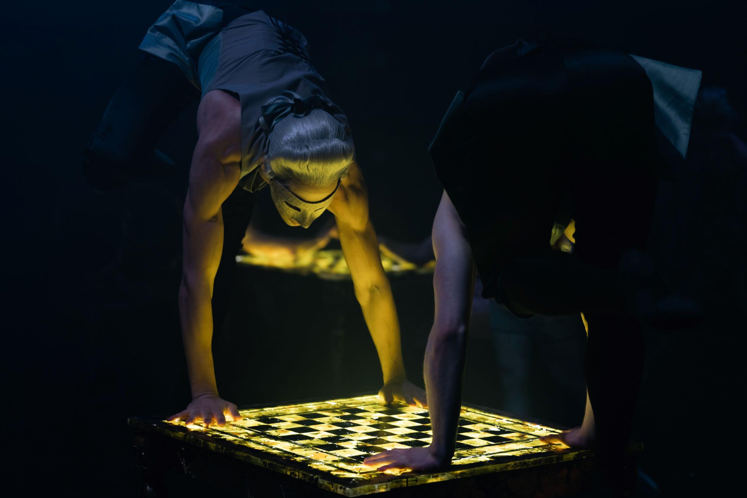 Two masked dancers look down at an illuminated chess board