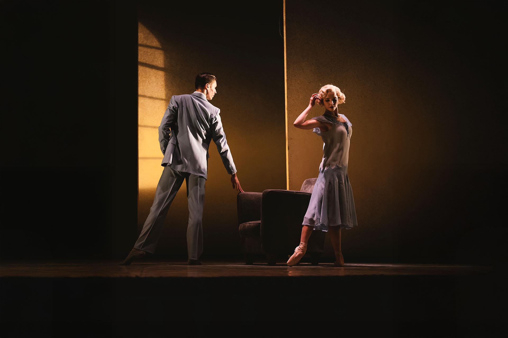 Dancers on stage in 1920 clothing, her a blue flapper dress, him is a sharp grey suit