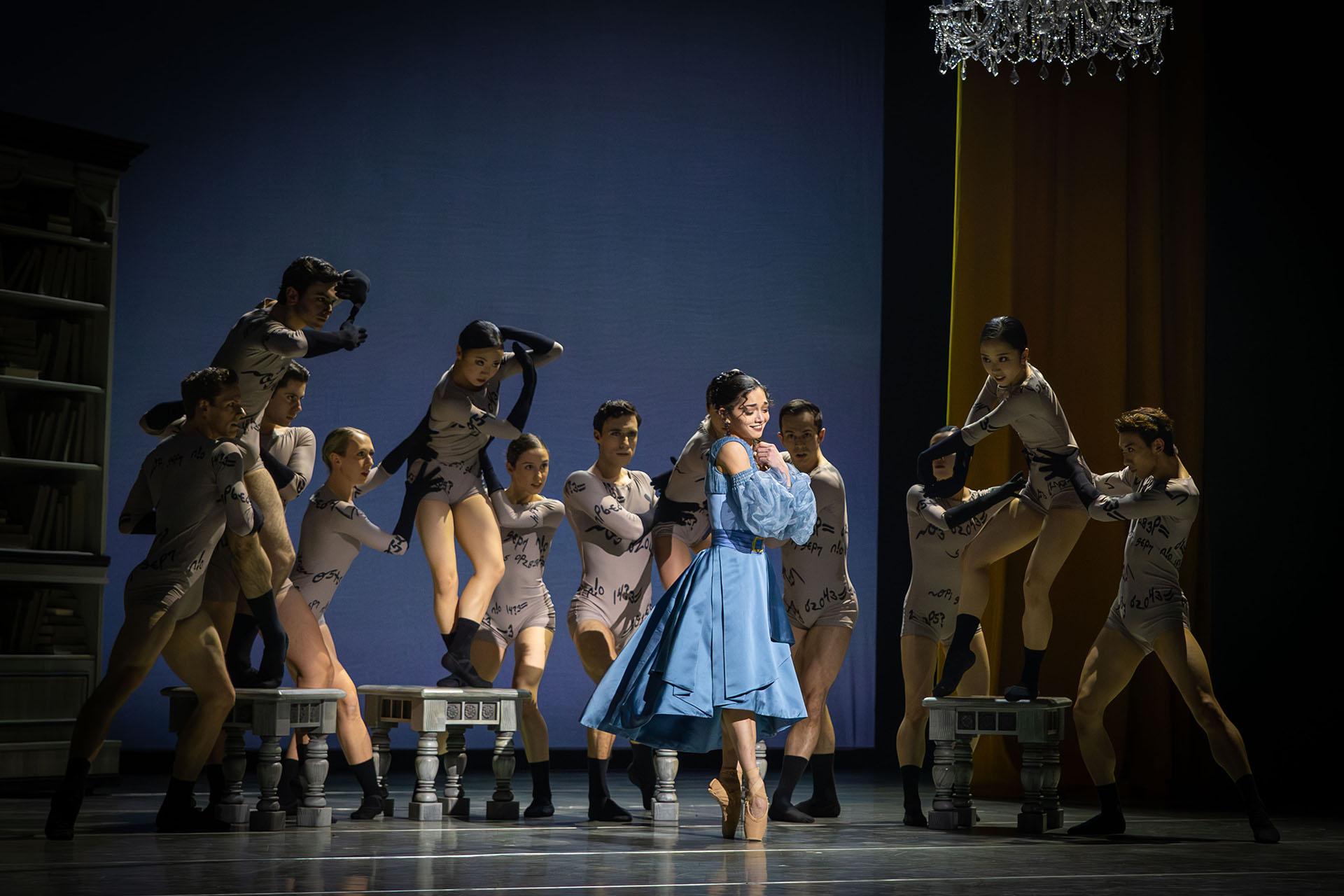 Woman in a blue dress smiles, smiling holding her hands to her chest, behind her dancers representing Anne Lister's beguiling words