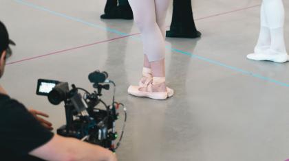 A camera operator in black focusing on a dancer in pointe shoes with legs crossed in a fifth position